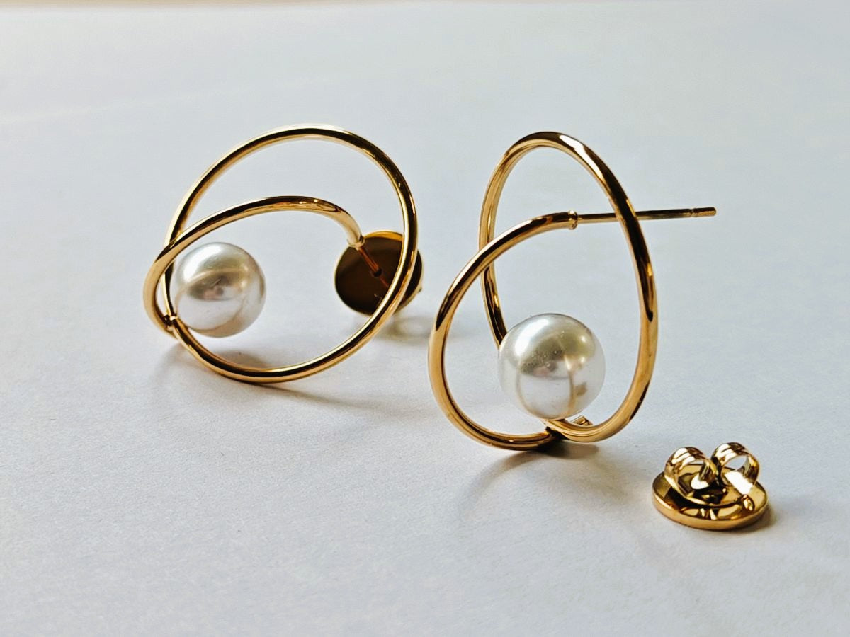 Detalle de los pendientes Nuvola mostrando el diseño tridimensional de las órbitas doradas y la perla blanca de acero waterproof baño en oro 18k con cierre de presión.